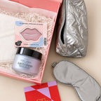 A curated beauty gift set arranged on a beige surface with a pink box filled with shredded paper. Visible items include a silver quilted zip pouch, a silver sleep mask, a pink lip mask package, and a jar labeled CeraVe Keratin Comfort, plus a red and pink gift card at the bottom.