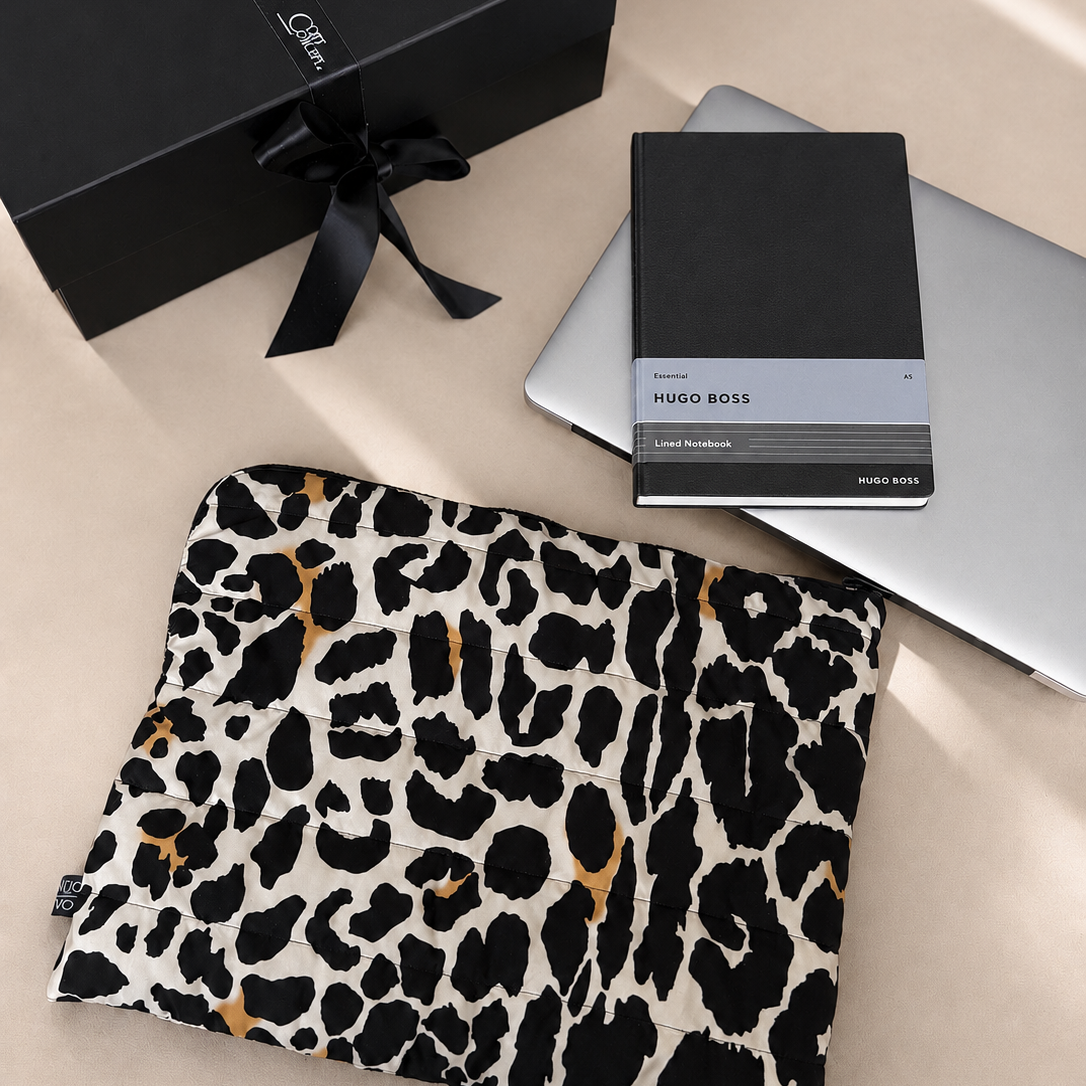 Leopard print pouch, black notebook, and silver laptop on a beige surface with a black gift box.
