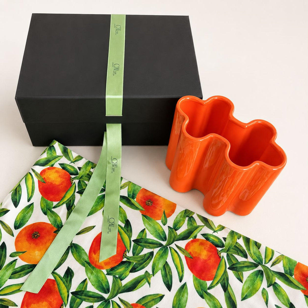 An orange ceramic flower-shaped vase displayed next to its black gift box and a green ribbon printed with the brand name 'O'Nile'. The gift box is wrapped in white paper featuring a green and orange citrus fruit pattern.