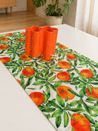 A white table runner printed with a green leaf and orange pattern, styled on a white table with a bright orange sculptural vase in the center.